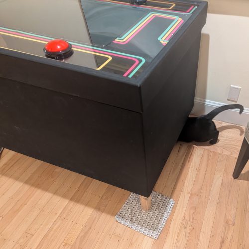 A black cat sticks her head under a giant black table. The sides of the table reach down just above the floor, so there's not a lot of room for her to squeeze under. There is a red button and some colorful lines on the surface of the table.